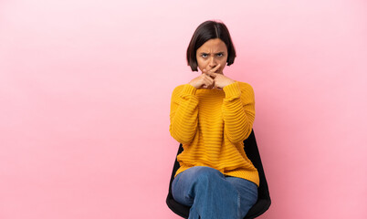 Young mixed race woman sitting on a chair isolated on pink background showing a sign of silence...