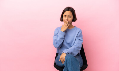 Young mixed race woman sitting on a chair isolated on pink background yawning and covering wide...
