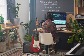 Rear view of young male musician in headphones sitting by his workplace in domestic room in front...