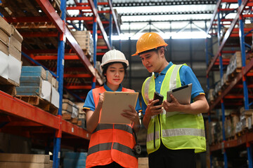 Two warehouse workers wearing safety hats are holding board and checking shipping stocks in...