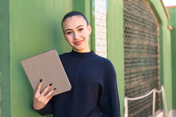 Young moroccan girl holding a tablet at outdoors smiling a lot