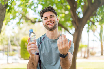 Young handsome sport man with a bottle of water at outdoors inviting to come with hand. Happy that...