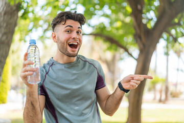 Fototapeta premium Young handsome sport man with a bottle of water at outdoors surprised and pointing finger to the side