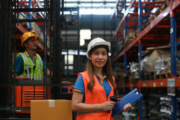 A confident Asian female warehouse worker working with bar code scanner while a man worker driving...