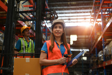 Portrait of a Confident Asian female worker holding a scanner and clipboard with a man working on a forklift as the background, Logistic distribution warehouse, Teamwork concept