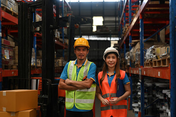 Portrait of a confident forklift truck with a male driver with arms crossed and a smiling female warehouse operator, stock delivery, or supply chain concept