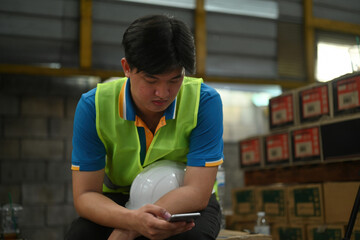 A warehouse worker male on break relaxes and reads the news on his smart phone in an industrial factory