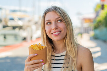 Young blonde woman holding fried chips at outdoors looking up while smiling