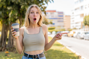 Young blonde woman holding a take away coffee at outdoors surprised and pointing side