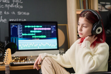 Cute restful girl with dyed red hair sitting in home studio against computer screen and listening...