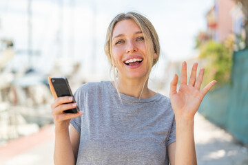 Young blonde woman using mobile phone at outdoors saluting with hand with happy expression