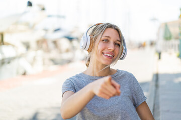 Young blonde woman at outdoors listening music and pointing to the front