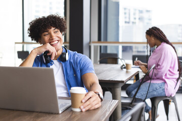 Biracial man with curly hair smiles, using laptop and holding a coffee cup