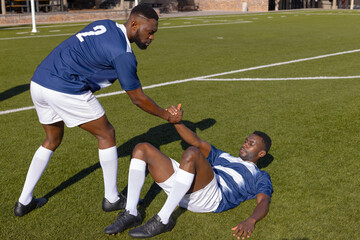 Two African American male athletes play rugby, one helping the other up