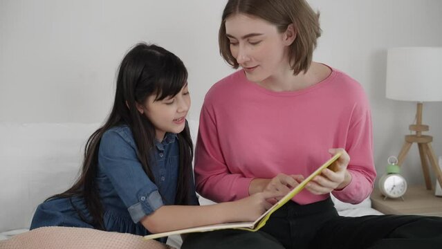 Caucasian mother reading story book to cute daughter listening story. Asian girl pointing while talking about story before bed time. Happy family sitting on bed while spend time together. Pedagogy.