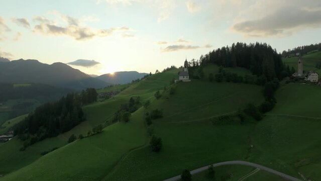 Aerial cinematic shot of lush green hills in La Val village, South Tyrol, Dolomites, Italy. Drone flies near a church, Sass de Putia mountain in backlit scene. LuPa Creative.