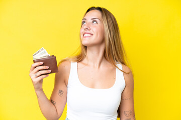 Young caucasian woman holding a wallet isolated on yellow background looking up while smiling