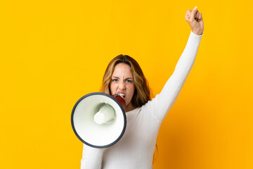 Young blonde woman isolated on yellow background shouting through a megaphone to announce something