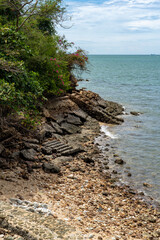 Steep rocky seashore with an old stone staircase, overgrown with tropical vegetation at low tide. Sky with clouds in the background.