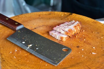 A knife is on a wooden cutting board with a piece of meat on it. The knife is sharp and has a black...
