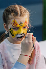 A woman professional artist paints with a brush, a colored pencil on the face of a child, a beautiful little girl, face painting, makeup, watercolor drawing at a party. Photography, creative process.
