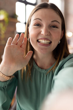 Young Caucasian professional woman in green top waves at camera on video call