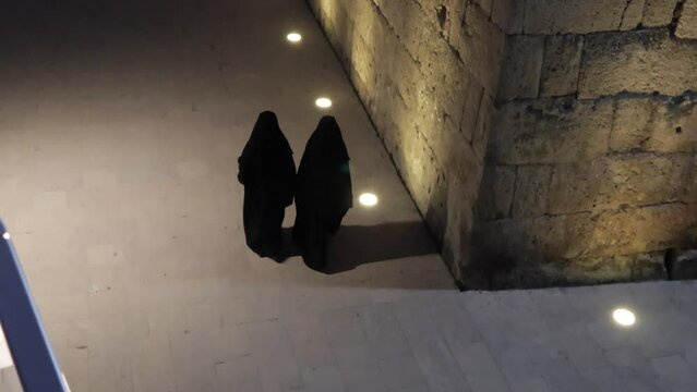 Traditional women's outerwear in Muslim countries. Women in black niqabs walk at night along a lighted street next to an ancient stone wall. Religious women wear a burqa. Muslim robes.