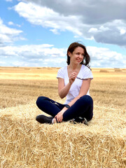 Happy young woman model sitting on a haystack, harvest time. Countryside girl on high haystack on the field.