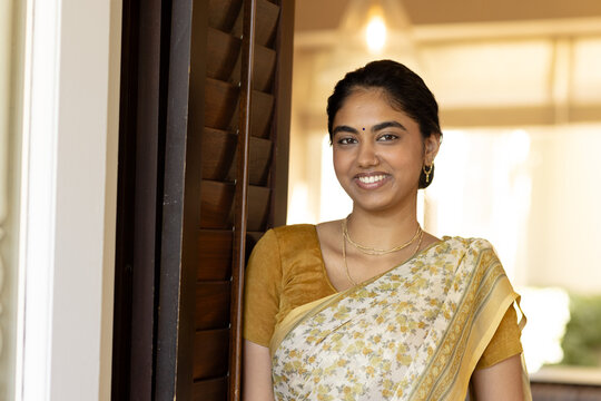 Indian young woman leaning against door frame at home, wearing traditional saree