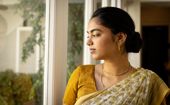 Indian young woman wearing traditional saree, looking out window at home