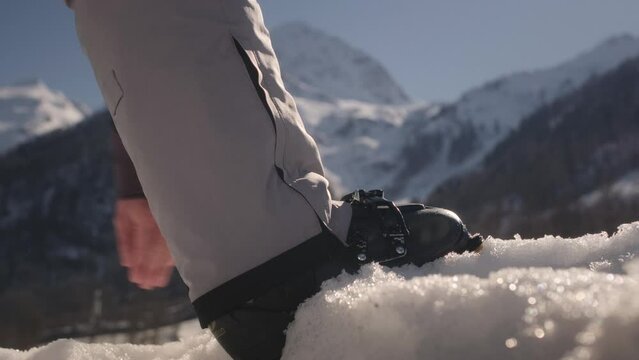 Skier puts his foot on the snow, fastens the clasp. Adjusting boat buckle on ski boot, close up. Alpine snowy mountains in sunny weather. Preparation for skiing, essential ski gear, winter sport