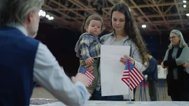 Woman with baby on hands talks with polling officer and takes bulletin. Female American citizen comes to vote in polling station. Political races of US presidential candidates. National Election Day.