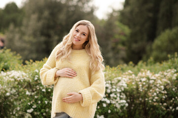 Happy pregnant woman 25-30 year old wearing yellow cozy knitted sweater posing over blooming flowers outdoor in park. Maternity time. Spring season.