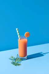 Jugo de zanahoria en un vaso transparente con pajita adornado con rodaja de zanahoria sobre fondo azul