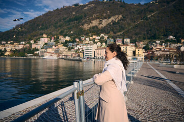 Happy young woman in the promenade, contemplating the lake of Come, relaxing outdoors over alpine mountains background, on a sunny winter day. People and beauty in nature. Italy. Travel destinations