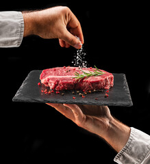 Chef is salting or seasoning raw ribeye steak laying on graphite serving board. Black background.