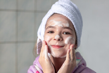 A girl in the bathroom with a white towel on her head washes her face with cleansing foam, smiles