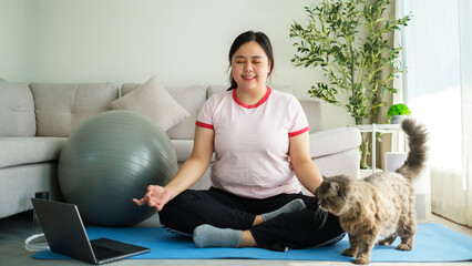 Young plus size woman practising yoga meditating on mat at cozy home. Wellness and healthy...