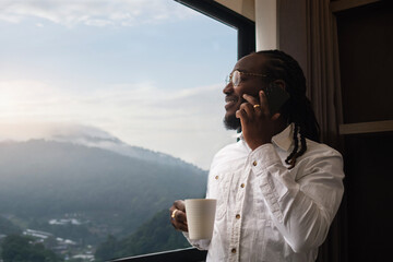 Smiling African American businessman with coffee cup standing next to the large windows having pleasant phone conversation and looking away