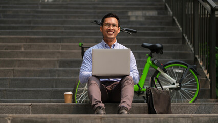 Smiling businessman using laptop on staircase in the city near his bicycle.