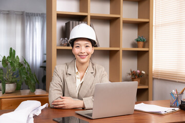 Asian architect or small business contractor female wearing hardhat looking camera using technology laptop everywhere working, engineer worker woman happy working at home office desk with a blueprint