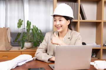 Asian architect or small business contractor female wearing hardhat looking camera using technology laptop everywhere working, engineer worker woman happy working at home office desk with a blueprint