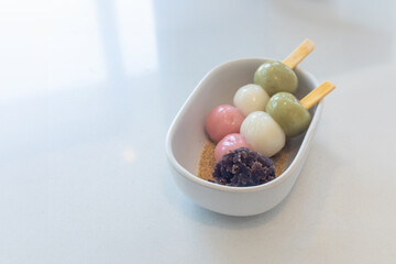 A bowl of assorted desserts, including green and pink balls and a brown dessert. The bowl is white and has a wooden stick in it