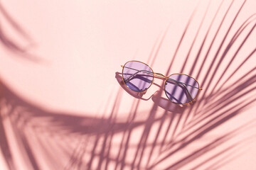 Blue glasses on a pink background with contrasting shadows of a palm tree. Summer vacation concept