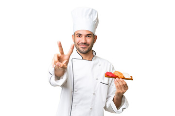 Young caucasian chef holding a sushi over isolated background smiling and showing victory sign