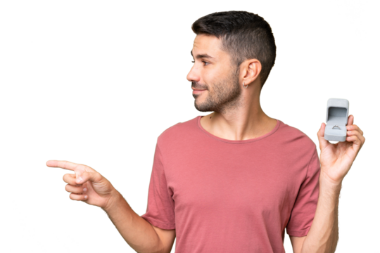 Young handsome caucasian man holding a engagement ring over isolated background pointing to the side to present a product - Powered by Adobe