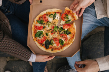 Top view image of couple grab slices of pizza from box at the outdoor. Man and woman hands taking pizza. Vegan pizza with fresh tomatoes basil and broccoli. Lactose and gluten free