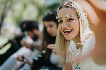 Cheerful young woman wearing a daisy chain headband laughs while enjoying a sunny day at a park picnic with friends. Soft focus on background.