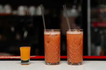 two glasses with red strawberry cocktails with straws and glass with orange juice on bar stand,...