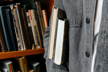 young man in grey jacket holds stack of books ander arm, bookshelves in background, student...